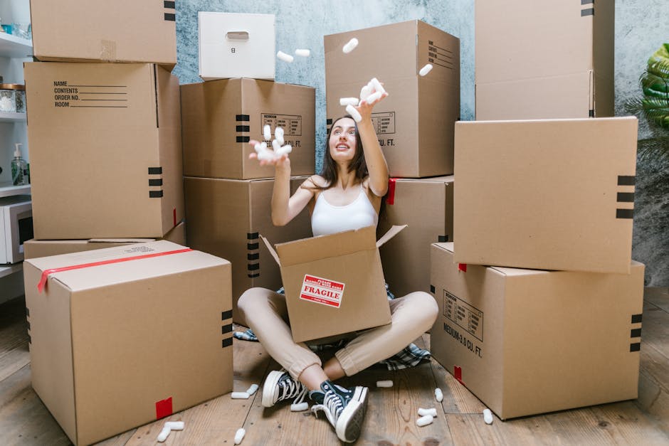 A young woman with long dark hair, dressed in a white tank top, beige pants, and sneakers, is sitting cross-legged on a wooden floor inside a room during a house removal process. She is smiling and to