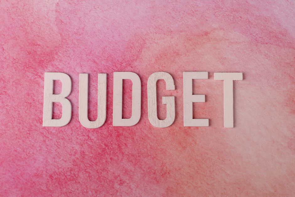 A young woman with long dark hair, dressed in a white tank top, beige pants, and sneakers, is sitting cross-legged on a wooden floor inside a room during a house removal process. She is smiling and to