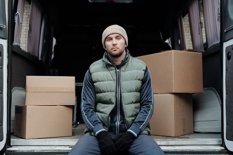 A man wearing a beige knit beanie, a green puffer vest, and a blue tracksuit sitting on the edge of an open moving van in front of a property during daylight. Inside the van, multiple cardboard boxes of various sizes, some sealed with packing tape, are stacked neatly on the floor, with additional boxes visible towards the back. The van's interior includes black walls and a metal floor, while the background shows the entrance to a house or flat with a doorway and exterior wall. The scene captures the process of home relocation, with the man possibly taking a break during furniture transport or packing and loading activities, supported by the presence of packing materials and the vehicle designated for removals. Man With a Van Bromley provides professional removal services, aligning with the context of this furniture transport and moving process.