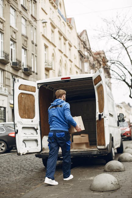 A man wearing a blue jacket, blue trousers, and white sneakers is seen loading cardboard boxes into the rear of a white cargo van parked on a cobbled street in Bromley. The van’s back doors are open, revealing an interior lined with plywood panels, ready for home relocation or furniture transport. The boxes vary in size and are made of cardboard, some wrapped with plastic for protection. The street is lined with multi-storey residential buildings with large windows and balconies. A few parked cars are visible along the pavement, and leafless trees indicate a colder season. The scene captures the loading process as part of packing and moving activities, with the man strategically placing boxes inside the van to optimise space while preparing for a house removal. The company, Man With a Van Bromley, provides professional removals and moving services in this urban environment, supporting efficient logistics for local relocations.
