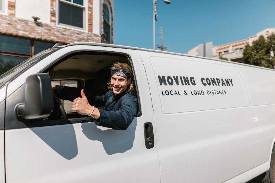 A man wearing a beige knit beanie, a green puffer vest, and a blue tracksuit sitting on the edge of an open moving van in front of a property during daylight. Inside the van, multiple cardboard boxes of various sizes, some sealed with packing tape, are stacked neatly on the floor, with additional boxes visible towards the back. The van's interior includes black walls and a metal floor, while the background shows the entrance to a house or flat with a doorway and exterior wall. The scene captures the process of home relocation, with the man possibly taking a break during furniture transport or packing and loading activities, supported by the presence of packing materials and the vehicle designated for removals. Man With a Van Bromley provides professional removal services, aligning with the context of this furniture transport and moving process.