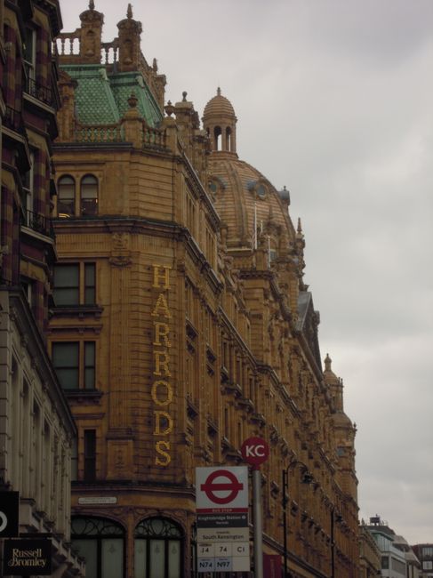 A detailed exterior view of a historic, ornate building with yellow brickwork and intricate architectural features, located in Bromley North. The building has a prominent vertical sign reading 'HARRODS' attached to its facade. In the foreground, there is a red and white London Underground bus stop sign with various bus route information below. The scene captures the urban environment during daytime with a cloudy sky overhead, and the building's architectural details include domed elements, turrets, and decorative finials, exemplifying classic city architecture. This setting aligns with the context of urban house removals, where detailed building surroundings and transportation options are relevant. Man With a Van Bromley provides professional moving and transport services in such city environments, assisting with the logistics of home relocation and furniture transport within this area.