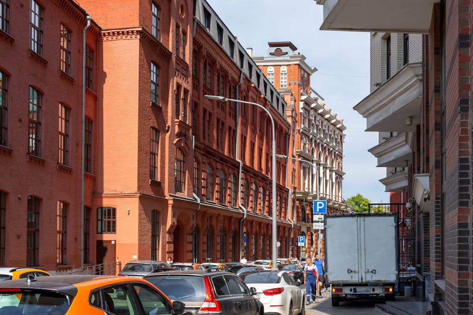 A narrow street in Bromley with multi-storey red brick buildings on both sides, featuring tall, arched and rectangular windows, some with window sills. Parked cars, including a yellow, silver, and white vehicles, line the curb along the street. A white delivery truck is parked on the right side, and a person in blue and white clothing is walking nearby. Several parking signs are visible, indicating parking restrictions, and a streetlight extends over the road. The scene is illuminated by daylight, highlighting the urban environment suitable for home relocation or furniture transport as part of professional removals services. The setting reflects a typical tight street environment where logistics and careful loading are essential for effective house removals, with Man With a Van Bromley operating in this area for efficient furniture transport and moving solutions.