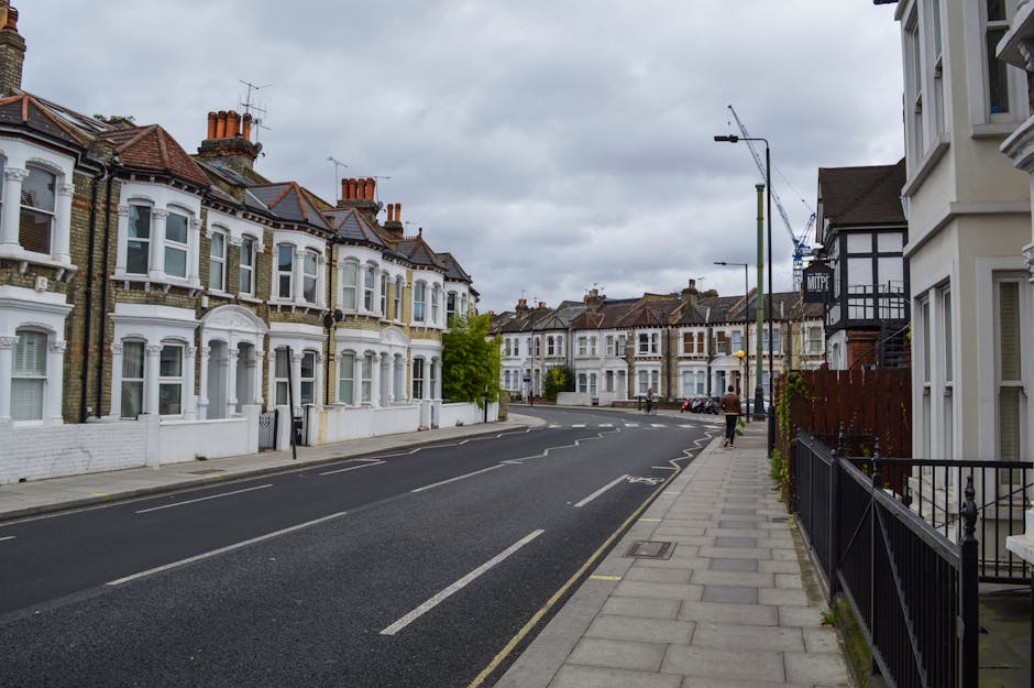 A wide residential street in Bromley with Victorian-style terraced houses featuring white facades, bay windows, and pitched roofs with red brick chimneys; the overcast sky casts diffuse light over the scene. The street has a paved sidewalk on the right side with black metal railings, and a few pedestrians are visible in the distance. The road is marked with dashed white lines and has a designated parking lane, although no vehicles are parked at the moment. There are street lamps, a few trees, and construction cranes visible further down the street, suggesting ongoing development. Inside the frame, part of a white van is visible, parked on the street, near an area where furniture and packing materials such as cardboard boxes and blankets could be loaded or unloaded during a house or flat relocation. The scene depicts a calm and tidy urban environment suitable for home relocation and furniture transport, with the presence of a local removals firm like Man With a Van Bromley implied in the context.