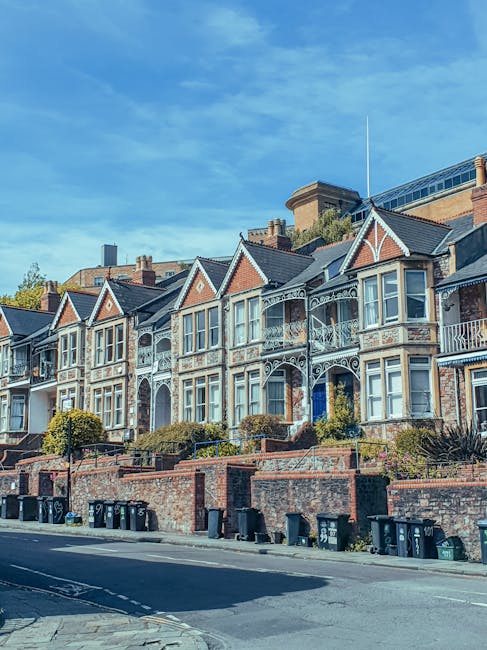 A row of historic Victorian-style terraced houses situated along a residential street in Bromley, featuring ornate white decorative trim, bay windows, and small front gardens with shrubs and trees. The houses are built with brickwork and have pitched roofs. In the foreground, there is a paved sidewalk with a line of black wheelie bins placed along the curb. The street appears calm and well-maintained, with clear weather and a blue sky overhead. The image captures the typical architecture associated with household relocations and property moving services, as provided by Man With a Van Bromley, with the focus on the external environment of residential properties ready for home relocation or furniture transport.